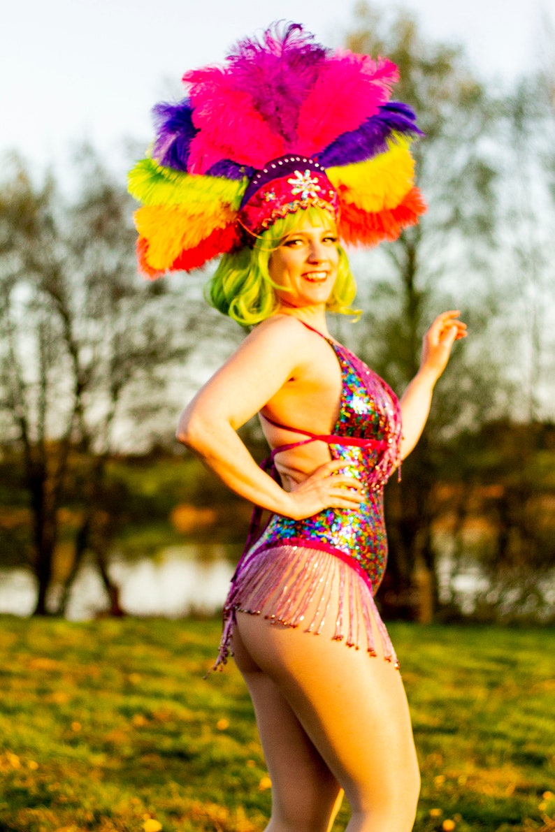 May include: A vibrant burlesque costume featuring a sequined bodysuit with fringe, a colorful feathered headdress, and green wig. The costume is predominantly pink, with accents of purple, yellow, and blue. The outfit is designed for stage performance.
