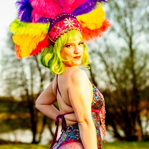 May include: A vibrant, colorful headdress with a rainbow of feathers, paired with a sequined bodysuit. The costume features a pink, purple, and blue color scheme, with beaded fringe. The model has green hair and a red lip.