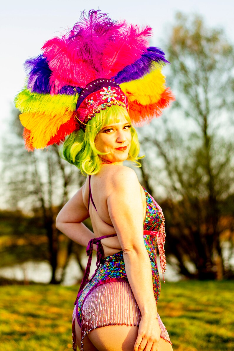 May include: A vibrant, colorful headdress with a rainbow of feathers, paired with a sequined bodysuit. The costume features a pink, purple, and blue color scheme, with beaded fringe. The model has green hair and a red lip.