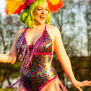May include: A vibrant, sequined bodysuit with fringe, in a rainbow of colors, is worn with a matching headdress of feathers. The headdress is pink, purple, yellow, and green. The person is smiling and laughing.