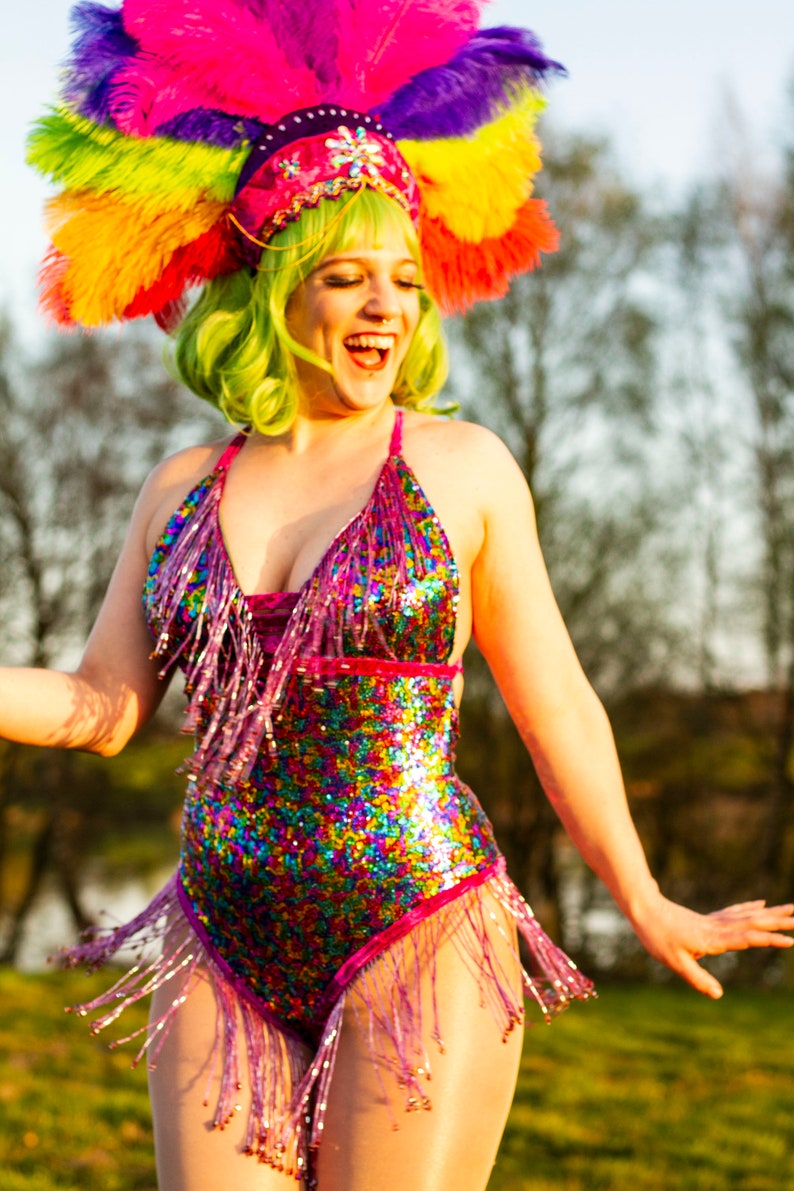 May include: A vibrant, sequined bodysuit with fringe, in a rainbow of colors, is worn with a matching headdress of feathers. The headdress is pink, purple, yellow, and green. The person is smiling and laughing.