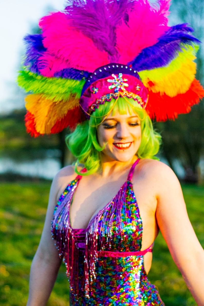May include: A vibrant, sequined halter top and matching headdress with a rainbow of feathers. The outfit features a pink, purple, and blue color scheme with fringe detailing. The model is wearing a green wig.
