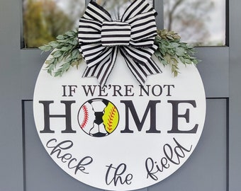 Puede incluir: Un letrero redondo de madera blanca con el texto "IF WE'RE NOT HOME check the field" en escritura negra. Un gráfico de béisbol y fútbol está en el centro. El letrero está decorado con un lazo a rayas blanco y negro y vegetación.