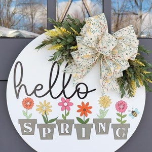 以下が含まれることがあります： 黒い筆記体で「hello spring」と書かれた白い丸い木製ドアハンガー。ハンガーは、花柄のリボン、緑の葉、カラフルな花が鉢植えになっている。
