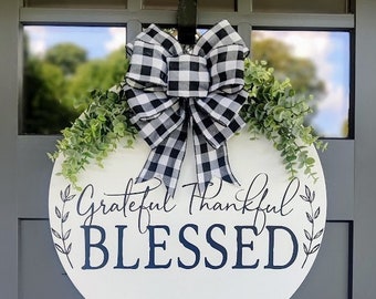 May include: A white round wooden door hanger with the text "Grateful Thankful Blessed" in black script. The hanger is decorated with a black and white checkered bow and greenery.