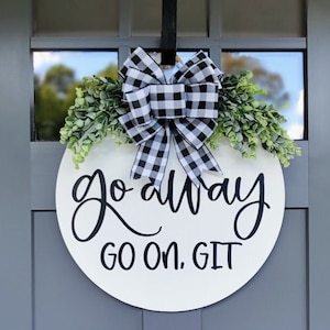 Könnte beinhalten: Ein rundes weißes Holzschild mit schwarzer Aufschrift "go away go on. git" hängt an einer grauen Tür mit einer schwarz-weißen karierten Schleife und Grün.
