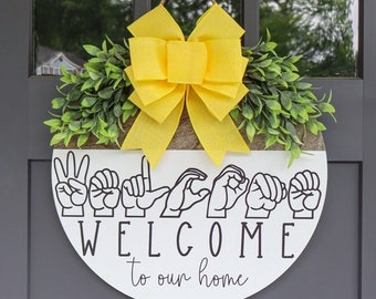 May include: A white round sign with the word "WELCOME" spelled out in sign language. The sign also says "to our home" in cursive writing. The sign is decorated with a yellow bow and green leaves.