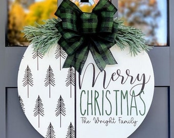 May include: A round white wooden door hanger with a black and white pine tree pattern on the left side. The right side has the text "Merry Christmas" in green script and "The Wright Family" in black script. A green and black plaid bow with green pine sprigs is attached to the top.