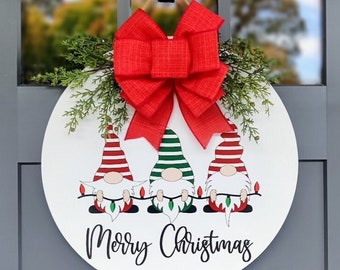May include: A white round door hanger with three gnomes holding string lights. The gnomes are wearing red and white striped hats, green and white striped hats, and red and white striped hats. The gnomes are holding string lights that are lit up. The door hanger has a red bow and green sprigs of greenery. The text "Merry Christmas" is written in black cursive on the door hanger.