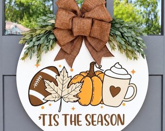 May include: A round white wooden door sign featuring a brown burlap bow and green foliage at the top. The graphic includes a brown football, a maple leaf, an orange pumpkin, and a mug with a heart. The text reads 'TIS THE SEASON in brown.