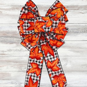 Puede incluir: Un lazo decorativo con un patrón de cuadros blancos y negros, adornado con hojas de otoño naranjas y rojas. El lazo tiene largas cintas, ideal para decoraciones otoñales. Las hojas tienen vetas detalladas.