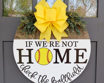 Puede incluir: Un colgador de puerta redondo de madera con fondo blanco y letras negras que dice "Si no estamos en casa, revisa el campo de béisbol". Una pelota de softball amarilla con costuras rojas está centrada en el letrero. Un lazo amarillo con hojas verdes está unido a la parte superior del letrero.