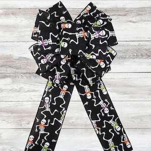 Può includere: Un grande fiocco nero con un motivo ripetuto di scheletri bianchi con papillon colorati. Il fiocco è fatto di un nastro largo ed è adatto per decorazioni di Halloween o a tema spettrale.