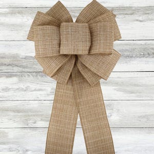 Puede incluir: Un lazo decorativo grande hecho de tela beige con un patrón de cuadros. El lazo presenta múltiples bucles y largas colas fluidas. La tela tiene una apariencia texturizada, y el lazo está sobre un fondo de madera blanca.