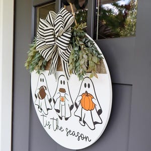 May include: A white round wooden door hanger with three ghosts holding a football, a coffee cup, and a pumpkin. The ghosts are white with black eyes and mouths. The text "Tis the season" is written in black cursive below the ghosts.