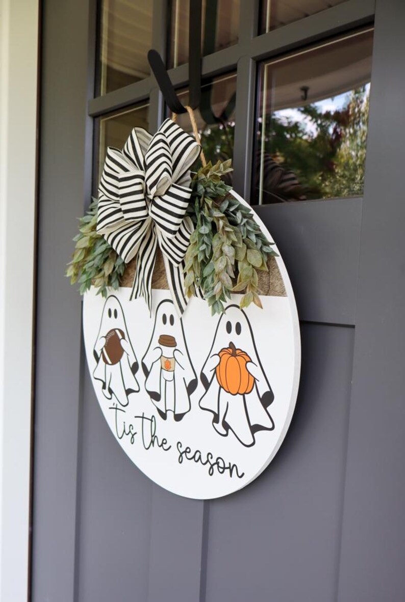 May include: A white round wooden door hanger with three ghosts holding a football, a coffee cup, and a pumpkin. The ghosts are white with black eyes and mouths. The text "Tis the season" is written in black cursive below the ghosts.