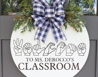 May include: A round white wooden classroom door sign with the words "WELCOME TO MS. DEROCCO'S CLASSROOM" in brown lettering. The sign features hand gestures spelling "WELCOME" in American Sign Language. It is decorated with greenery and a blue and white checkered bow.
