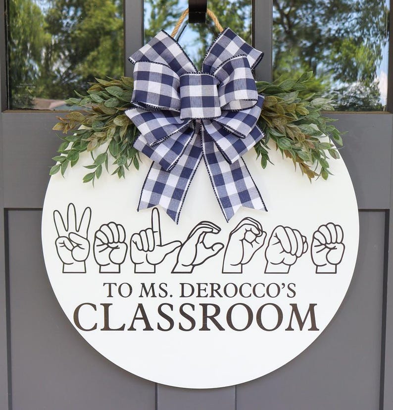 Pu&ograve; includere: Un cartello rotondo in legno bianco per la porta della classe con le parole "WELCOME TO MS. DEROCCO'S CLASSROOM" in lettere marroni. Il cartello presenta gesti delle mani che compongono "WELCOME" nella lingua dei segni americana. &Egrave; decorato con verde e un fiocco a scacchi blu e bianco.