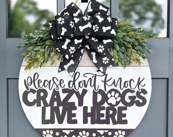 Könnte beinhalten: Rundes Holzschild mit dem Text "Please don't knock CRAZY DOGS LIVE HERE". Es hat einen schwarz-weißen Pfotenabdruckrand, einen grünen Blätterkranz und eine schwarze Schleife mit weißen Pfotenabdrücken und Knochen.