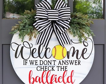 Puede incluir: Un letrero de bienvenida redondo de madera con temática de softbol. El letrero presenta la palabra "Welcome" en escritura negra, una pelota de softbol amarilla y el texto "If we don't answer check the ballfield" en rojo. Está decorado con vegetación y un lazo a rayas blanco y negro.