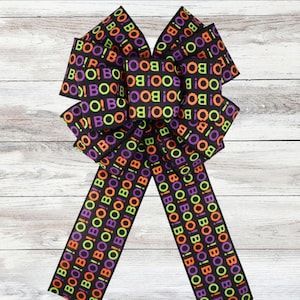 Può includere: Un fiocco decorativo di Halloween realizzato con nastro nero. Il nastro è stampato con la parola "BOO!" in arancione, verde e viola. Il fiocco ha più anelli e lunghe code, perfetto per decorazioni festive.