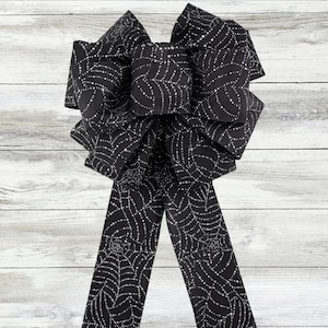 Può includere: Un grande fiocco nero con un motivo a ragnatela bianca. Il fiocco ha più anelli e lunghe code, perfetto per decorare. Il tessuto ha una lucentezza sottile. Ideale per Halloween o decorazioni a tema spettrale.