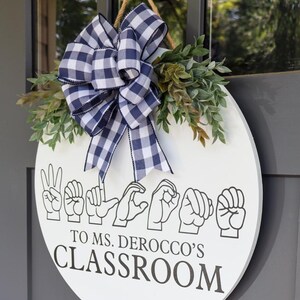Pu&ograve; includere: Un cartello rotondo bianco per la classe con le parole "Benvenuti nella classe della signora Derocco" in linguaggio dei segni. Decorato con un fiocco a scacchi blu e bianco e vegetazione.