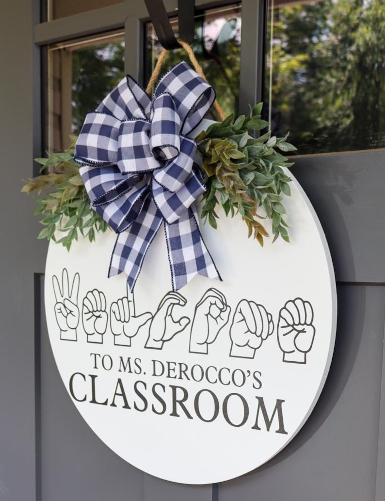 Pu&ograve; includere: Un cartello rotondo bianco per la classe con le parole "Benvenuti nella classe della signora Derocco" in linguaggio dei segni. Decorato con un fiocco a scacchi blu e bianco e vegetazione.