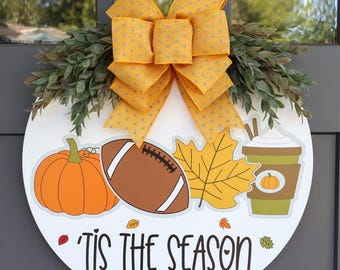 May include: A round white wooden door sign with a yellow bow and greenery. The sign features fall-themed graphics, including a pumpkin, football, leaf, and coffee cup. The text "'Tis the Season" is displayed in black script.