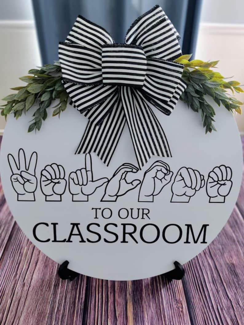 Pu&ograve; includere: Un cartello rotondo bianco per la classe con testo nero e un fiocco a righe bianche e nere. Il cartello recita "Benvenuti nella nostra classe" sia in linguaggio dei segni che in inglese. Il verde adorna la parte superiore del cartello.