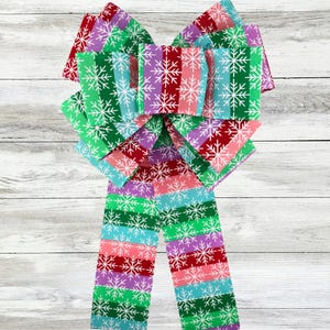 May include: A large, colourful bow with a striped pattern in red, green, purple, and blue. White snowflakes are printed on the stripes. The bow is made of fabric and is likely for holiday or seasonal decor.