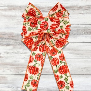 May include: A decorative bow featuring a repeating pattern of orange pumpkins and green vines on an off-white background. The bow has multiple loops and long tails, with orange trim. Perfect for fall or autumn decor.
