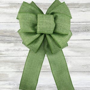 Puede incluir: Un lazo decorativo grande en un tono verde vibrante. El lazo presenta múltiples bucles y largas colas fluidas, hechas de una tela texturizada. El lazo está diseñado para la decoración y añade un toque de elegancia a cualquier entorno.