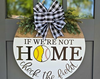 Puede incluir: Cartel de puerta redondo de madera con el texto "IF WE'RE NOT HOME check the field". El cartel presenta un gráfico de softbol y un lazo a cuadros en blanco y negro. La vegetación acentúa la parte superior.