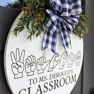 Pu&ograve; includere: Un cartello rotondo bianco per l'aula con testo nero e gesti delle mani che compongono "Benvenuti". Il cartello &egrave; decorato con verde e un fiocco a scacchi blu navy e bianco. Il testo recita "To Ms. Derocco's Classroom".