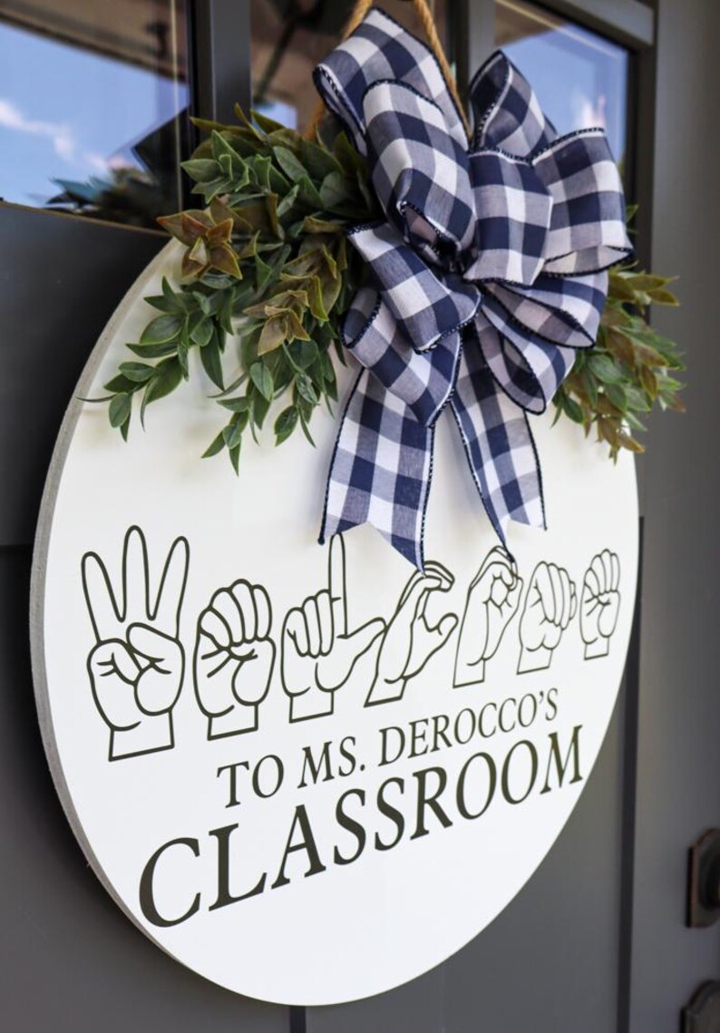 Pu&ograve; includere: Un cartello rotondo bianco per l'aula con testo nero e gesti delle mani che compongono "Benvenuti". Il cartello &egrave; decorato con verde e un fiocco a scacchi blu navy e bianco. Il testo recita "To Ms. Derocco's Classroom".