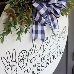 Pu&ograve; includere: Un cartello rotondo bianco per l'aula con testo nero e gesti della lingua dei segni italiana. Il cartello &egrave; decorato con un fiocco a quadri blu navy e bianco e fogliame verde. Il testo recita "TO MS. DEROCCO'S CLASSROOM."