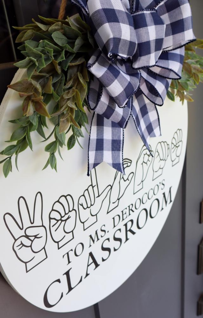 Pu&ograve; includere: Un cartello rotondo bianco per l'aula con testo nero e gesti della lingua dei segni italiana. Il cartello &egrave; decorato con un fiocco a quadri blu navy e bianco e fogliame verde. Il testo recita "TO MS. DEROCCO'S CLASSROOM."