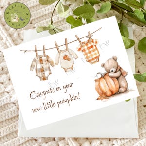 Pode incluir: Um cartão com uma ilustração em aquarela. O cartão apresenta roupas de bebê penduradas em um varal, um ursinho de pelúcia abraçando uma abóbora e o texto "Congrats on your new little pumpkin!". O cartão está em um envelope branco.