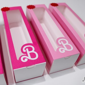 Puede incluir: Cuatro cajas rosas con ventanas transparentes y tapas blancas. Cada caja tiene una letra "B" blanca con un diseño de espiral decorativo. Las cajas están alineadas en una fila.