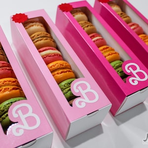 Può includere: Quattro scatole rosa con finestre trasparenti che mostrano macarons colorati. Ogni scatola ha una lettera "B" bianca sul davanti. Le scatole sono disposte in fila da sinistra a destra.