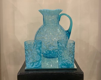 May include: A set of blue glass pitcher and two tumblers with a floral pattern. The pitcher has a wide mouth and a handle. The tumblers are short and have a straight side. The set is on a black pedestal.