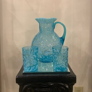 May include: A set of blue glass pitcher and two tumblers with a floral pattern. The pitcher has a wide mouth and a handle. The tumblers are short and have a straight side. The set is on a black pedestal.