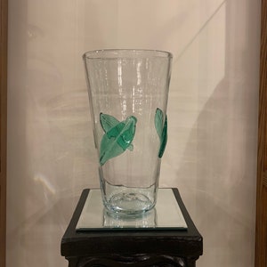 May include: A clear glass vase with a textured surface and green glass leaves attached to the side. The vase is sitting on a black pedestal.