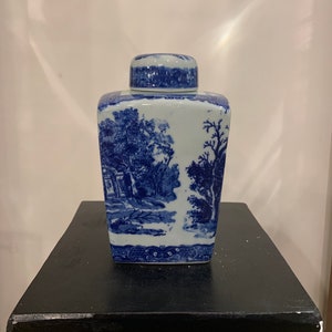 May include: A square blue and white porcelain jar with a lid. The jar features a blue and white landscape design with trees and a building.