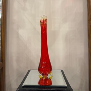 May include: A tall, red glass vase with a flared top and a yellow rim. The vase has a unique, textured design and stands on a small, mirrored pedestal.
