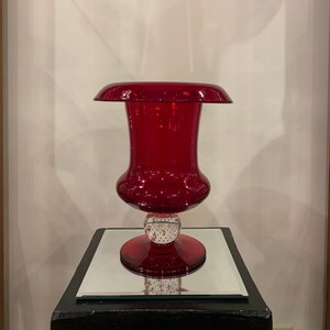 May include: A deep red glass vase with a wide, flared top and a round, textured base. The vase is on a black pedestal with a mirrored top.