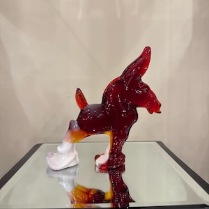 May include: A red glass donkey figurine with white accents. The donkey is standing on a mirrored surface.