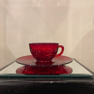 May include: A red glass teacup and saucer set with a decorative pattern. The cup and saucer are on a black surface with a mirrored top.