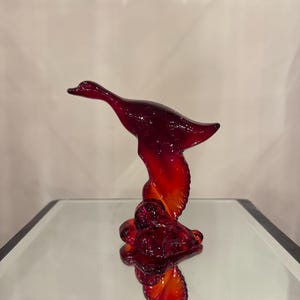 May include: A ruby red glass sculpture of a duck in flight. The duck has a long neck and outstretched wings, with a detailed base. The glass has a gradient of colors, transitioning from a deep red to a vibrant orange.